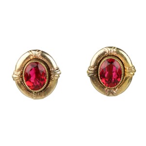 May include: A pair of gold-toned stud earrings. Each earring features a vibrant red oval-shaped gemstone set within a decorative gold frame. The earrings have a classic, elegant design, suitable for various occasions.