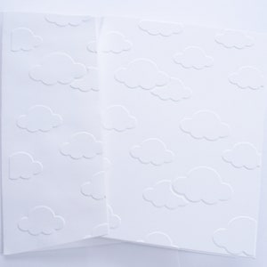 Może przedstawiać: Biały karton z wytłoczonym wzorem chmur. Projekt przedstawia puszyste chmury rozrzucone po powierzchni. Karta jest złożona, odsłaniając wnętrze. Idealny do artykułów papierniczych, zaproszeń lub projektów rzemieślniczych.
