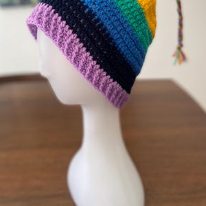 May include: A vibrant, hand-crocheted hat in a rainbow design, displayed on a white mannequin head. The hat features stripes of purple, navy, blue, green, yellow, and red. A small tassel hangs from the top. A fun and colourful accessory.