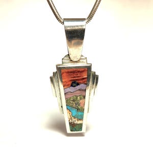 May include: Silver pendant necklace with a rectangular, multi-colored inlay depicting a landscape scene. The inlay features shades of red, purple, green, and blue. The pendant is suspended from a silver chain.