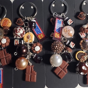 Puede incluir: Cuatro llaveros con aros plateados, cada uno adornado con dijes de barras de chocolate, caramelos y cuentas. Los dijes incluyen barras Snickers, botellas de Nutella y adornos con temática de chocolate. Los llaveros están dispuestos sobre un fondo oscuro.