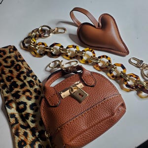 May include: An assortment of fashion accessories, including a small brown purse with a gold clasp, a brown heart-shaped charm, and two chain straps with tortoise shell links. A leopard print scarf is also included.