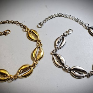 Puede incluir: Dos pulseras de conchas, una dorada y otra plateada, con un diseño de eslabones de cadena. Las pulseras están hechas de metal y presentan una serie de abalorios en forma de concha.