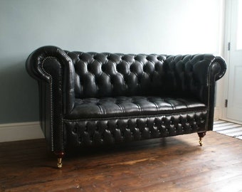 May include: A black leather tufted sofa with a button-tufted design. The sofa has a classic style and is upholstered in black leather. The sofa has four legs and is sitting on a wooden floor.