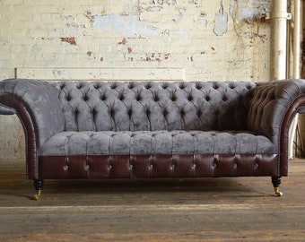 May include: A gray tufted sofa with brown leather trim and wooden legs. The sofa is in front of a white brick wall.