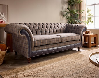 May include: A tufted, two-cushion sofa with a plaid pattern in brown, tan, and gray. The sofa has rolled arms and dark blue leather trim. The sofa sits on wooden legs and is placed on a woven rug. The room has a neutral color palette.