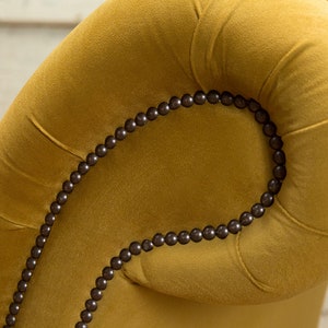 Puede incluir: Primer plano de un sill&oacute;n de terciopelo amarillo mostaza con una fila de cuentas marrones oscuras que delinean la parte superior del reposabrazos.