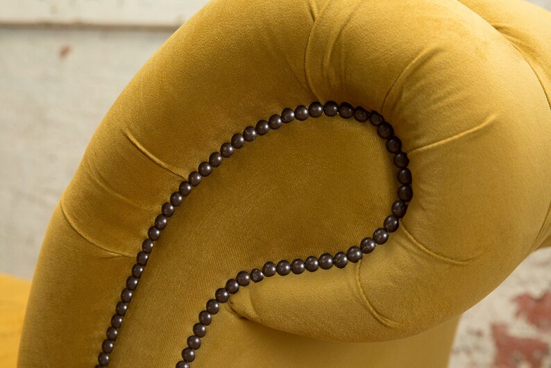 Puede incluir: Primer plano de un sill&oacute;n de terciopelo amarillo mostaza con una fila de cuentas marrones oscuras que delinean la parte superior del reposabrazos.