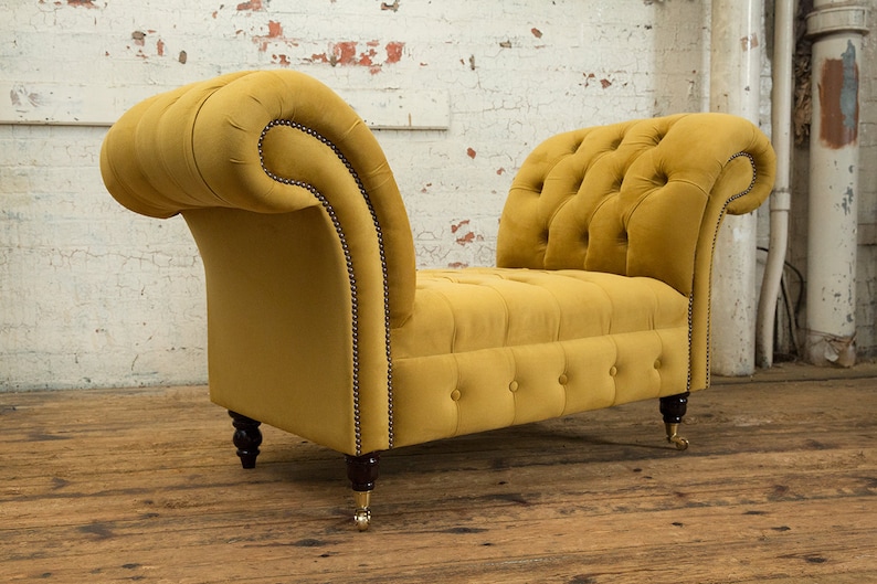 Puede incluir: Un banco tapizado en terciopelo amarillo mostaza con patas de madera y ruedas de lat&oacute;n. El banco tiene un respaldo y brazos curvos.