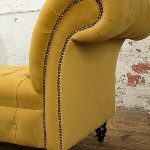 Puede incluir: Una chaise longue de terciopelo amarillo mostaza con respaldo capiton&eacute; y un brazo enrollado. La chaise longue tiene patas de madera marr&oacute;n oscuro con ruedas de lat&oacute;n.