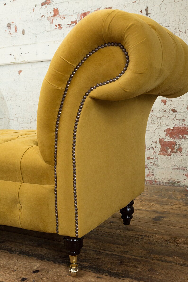 Puede incluir: Una chaise longue de terciopelo amarillo mostaza con respaldo capiton&eacute; y un brazo enrollado. La chaise longue tiene patas de madera marr&oacute;n oscuro con ruedas de lat&oacute;n.