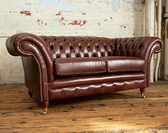 May include: A brown leather chesterfield sofa with button tufting and wooden legs. The sofa is in front of a white brick wall.