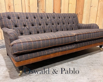 May include: A brown and gray plaid sofa with a tufted back and brown leather trim. The sofa has wooden legs with brass casters and is sitting on a concrete floor. The text "Oswald & Pablo" is visible in the foreground.