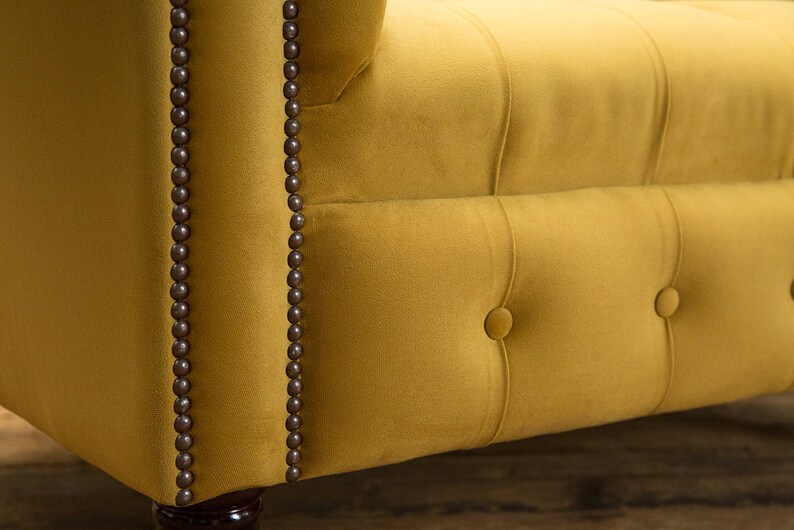 Puede incluir: Primer plano de un sof&aacute; de terciopelo amarillo mostaza con botones capitonn&eacute;s y ribetes marrones. El sof&aacute; tiene un dise&ntilde;o cl&aacute;sico con respaldo y brazos enrollados.
