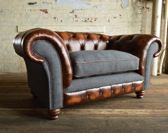 May include: A brown leather armchair with a gray fabric cushion. The armchair has a tufted back and a rolled armrest. The armchair is sitting on a wooden floor.