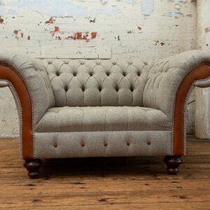 May include: A gray tufted armchair with brown leather accents and wooden legs. The chair is in front of a white brick wall.
