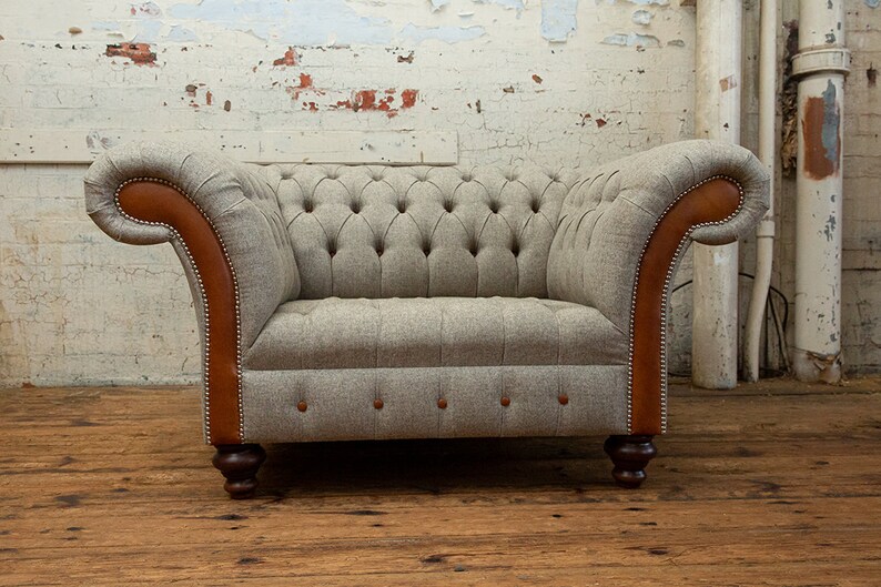 May include: A gray tufted armchair with brown leather accents and wooden legs. The chair is in front of a white brick wall.