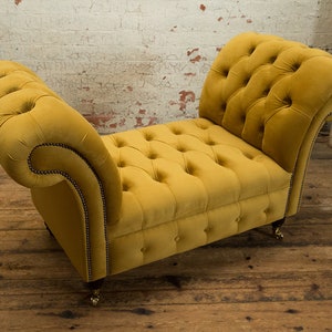 Puede incluir: Un banco de terciopelo amarillo con botones y brazos enrollados y patas de madera. El banco est&aacute; sobre un suelo de madera.