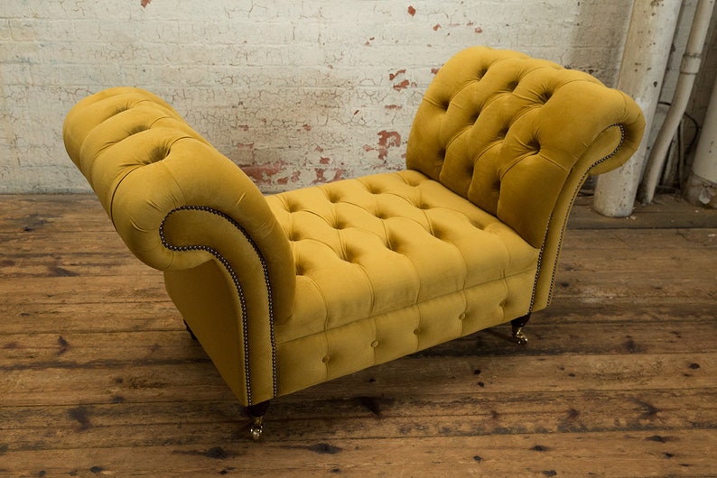 Puede incluir: Un banco de terciopelo amarillo con botones y brazos enrollados y patas de madera. El banco est&aacute; sobre un suelo de madera.