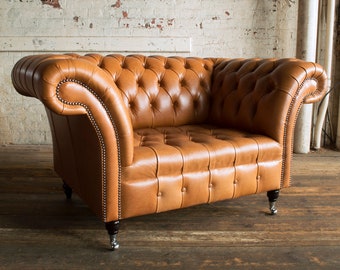 May include: A tufted, tan leather armchair with rolled arms and a low back. The chair features a button-tufted design and is trimmed with small silver studs. The chair sits on four short legs with silver caster wheels.