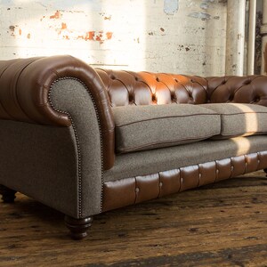 May include: A brown leather and tweed sofa with button tufting and wooden legs. The sofa is in a rustic setting with a white brick wall in the background.