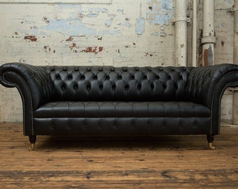 May include: A black leather chesterfield sofa with button tufting and rolled arms. The sofa is sitting on a wooden floor in front of a white brick wall.