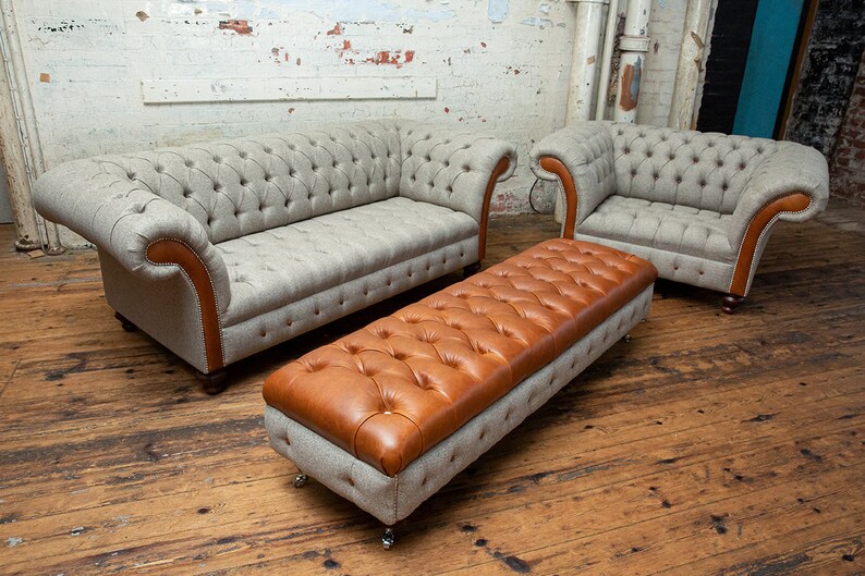 May include: A set of three tufted furniture pieces in a living room setting. A light gray sofa with brown trim, a matching armchair, and a brown leather tufted ottoman with a gray base.