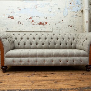 May include: A gray tufted sofa with brown leather trim and wooden legs. The sofa is in front of a white brick wall.
