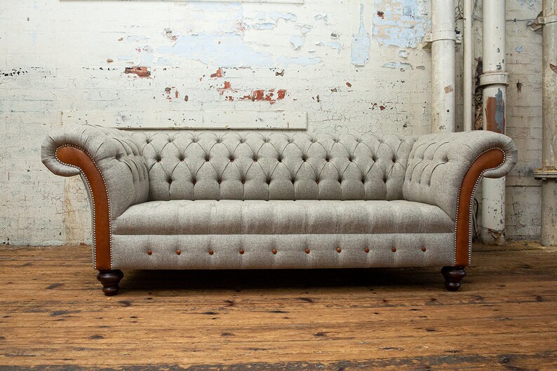May include: A gray tufted sofa with brown leather trim and wooden legs. The sofa is in front of a white brick wall.