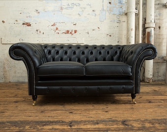 May include: A black leather chesterfield sofa with button tufting and rolled arms. The sofa is sitting on a wooden floor in front of a white brick wall.