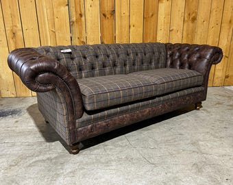 May include: A brown and plaid upholstered sofa with rolled arms. The sofa features a button-tufted back and leather accents. The cushions have a plaid pattern with brown, tan, and gold tones. The sofa sits on short, wooden legs.