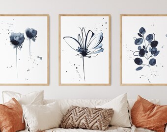 May include: Three framed watercolor prints featuring botanical designs in shades of blue and white. The artwork includes floral and leaf motifs with a minimalist aesthetic. The prints are displayed above a white sofa with orange and cream-colored pillows.