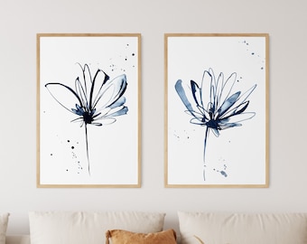 May include: Two framed floral art prints. Each print features a blue watercolor flower with delicate lines and ink splatters on a white background. The frames are a light brown color. The artwork is displayed above a beige sofa.