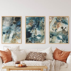 K&ouml;nnte beinhalten: Drei gerahmte abstrakte Aquarelldrucke. Das Kunstwerk zeigt eine Mischung aus Blau-, T&uuml;rkis- und Braunt&ouml;nen mit botanischen und strukturierten Elementen. Die Drucke werden &uuml;ber einem wei&szlig;en Sofa mit orangefarbenen und neutralfarbenen Kissen pr&auml;sentiert.