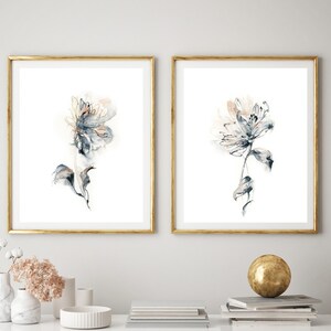 Puede incluir: Dos impresiones art&iacute;sticas florales enmarcadas. Cada impresi&oacute;n presenta una flor de acuarela en tonos azules, grises y melocot&oacute;n sobre un fondo blanco. Los marcos son dorados. Las impresiones se exhiben sobre una mesa blanca con objetos decorativos.