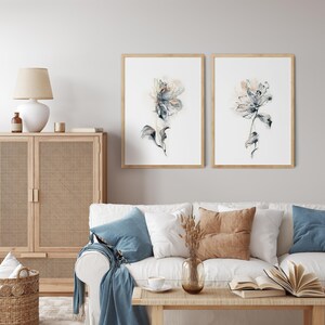 Puede incluir: Dos impresiones enmarcadas de acuarelas florales. Cada impresi&oacute;n presenta una flor delicada y abstracta en tonos azules, grises y beige sobre un fondo blanco. Los marcos son de madera clara. Las impresiones se exhiben sobre un sof&aacute; blanco con cojines azules y marrones.