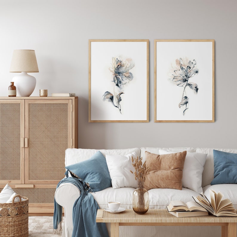 Puede incluir: Dos impresiones enmarcadas de acuarelas florales. Cada impresi&oacute;n presenta una flor delicada y abstracta en tonos azules, grises y beige sobre un fondo blanco. Los marcos son de madera clara. Las impresiones se exhiben sobre un sof&aacute; blanco con cojines azules y marrones.