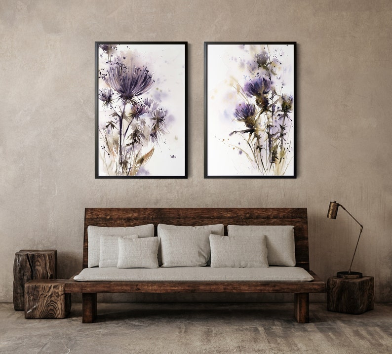 May include: Two framed watercolor paintings of purple thistle flowers. The artwork features a soft, blurred background with splashes of color. The frames are black, and the overall style is botanical and artistic.