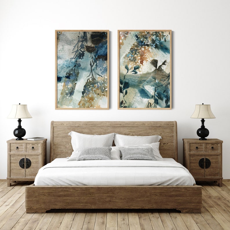 May include: Two framed watercolor paintings with abstract botanical designs in shades of blue, brown, and white hang above a wooden bed. The bedroom features matching wooden nightstands with black lamps and a white wall.