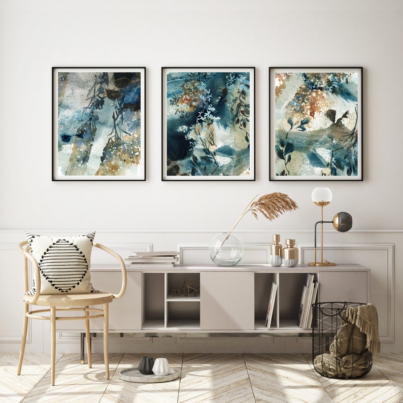 K&ouml;nnte beinhalten: Drei gerahmte abstrakte Aquarellmalereien in Blau-, T&uuml;rkis- und Braunt&ouml;nen. Das Kunstwerk zeigt botanische Elemente und wird &uuml;ber einer modernen Konsole mit dekorativen Gegenst&auml;nden ausgestellt.