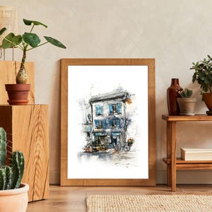 May include: A framed watercolour print of a European shopfront with blue shutters and a bicycle. The artwork is in a wooden frame, displayed with potted plants and a small wooden table. The decor has a natural, earthy aesthetic.