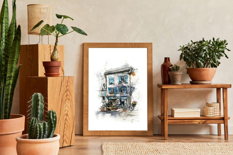 May include: A framed watercolour print of a European shopfront with blue shutters and a bicycle. The artwork is in a wooden frame, displayed with potted plants and a small wooden table. The decor has a natural, earthy aesthetic.