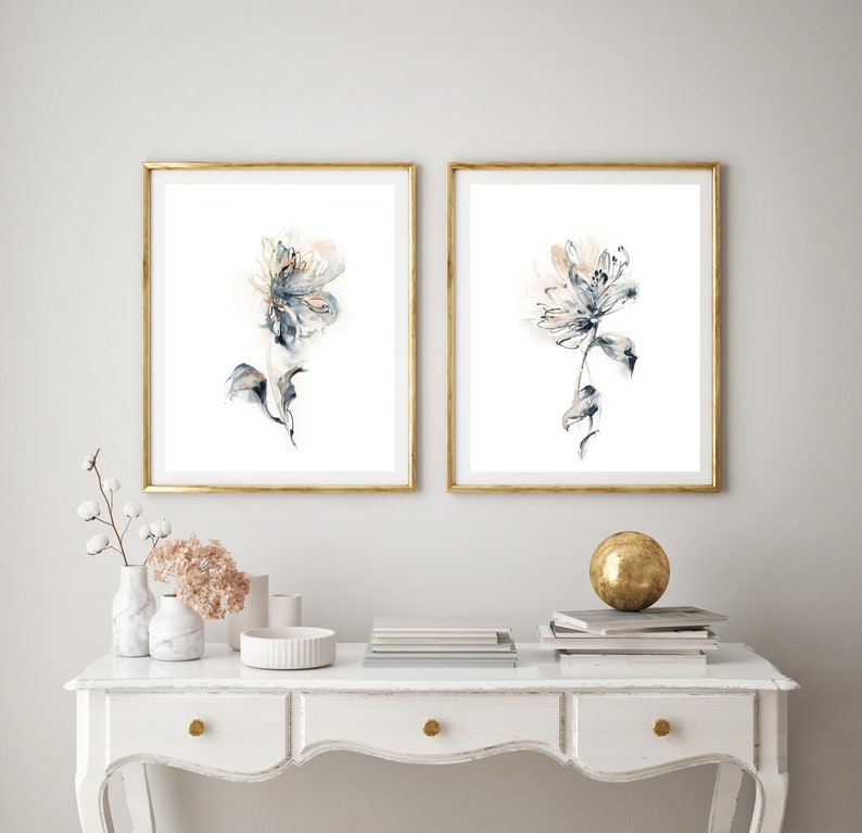 Puede incluir: Dos impresiones art&iacute;sticas florales enmarcadas. Cada impresi&oacute;n presenta una flor de acuarela en tonos azules, grises y melocot&oacute;n sobre un fondo blanco. Los marcos son dorados. Las impresiones se exhiben sobre una mesa blanca con objetos decorativos.