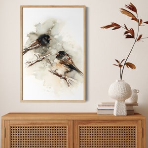 May include: Watercolour painting of two birds perched on a branch, set against a soft, neutral background. The birds feature black and brown plumage with touches of red. The artwork is framed in a light wood frame, adding a natural touch to the piece.