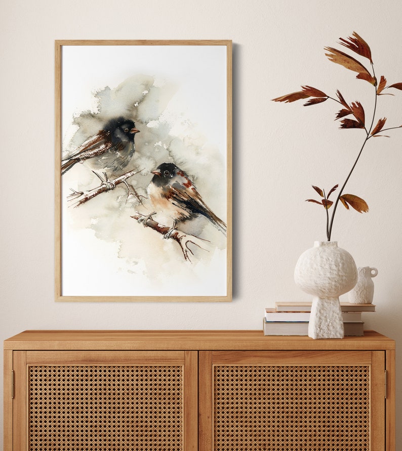 May include: Watercolour painting of two birds perched on a branch, set against a soft, neutral background. The birds feature black and brown plumage with touches of red. The artwork is framed in a light wood frame, adding a natural touch to the piece.
