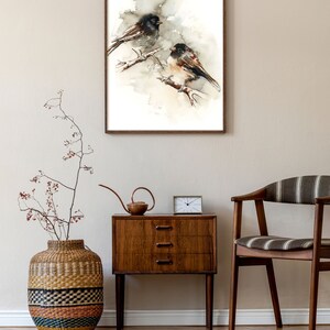 May include: A framed watercolour painting of two birds on a branch, in a neutral palette of browns and greys. It hangs above a wooden bedside table with a small clock and a decorative watering can. The room has a wooden floor.