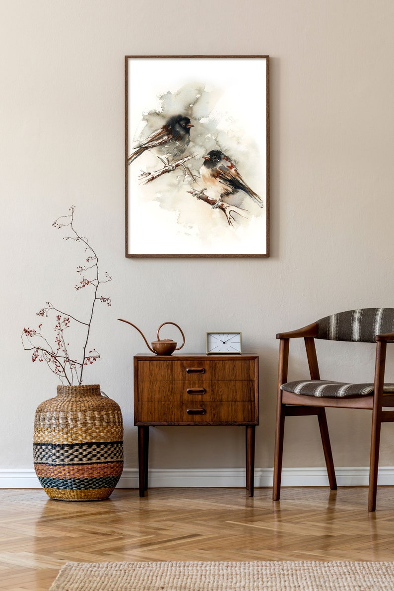 May include: A framed watercolour painting of two birds on a branch, in a neutral palette of browns and greys. It hangs above a wooden bedside table with a small clock and a decorative watering can. The room has a wooden floor.