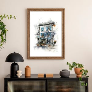 May include: Watercolour painting of a European building with blue shutters and a bicycle. The artwork is framed in a wooden frame and hangs on a cream wall. The scene is part of a home decor arrangement.