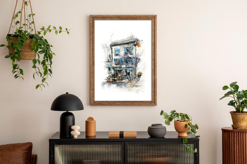 May include: Watercolour painting of a European building with blue shutters and a bicycle. The artwork is framed in a wooden frame and hangs on a cream wall. The scene is part of a home decor arrangement.