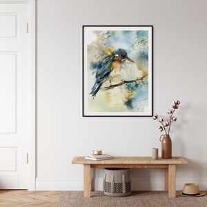 May include: Watercolor painting of a bird perched on a branch, framed in black. The bird features blue, yellow, and brown hues. The background has soft, blended colors of blue, yellow, and beige. The artwork is displayed on a white wall.